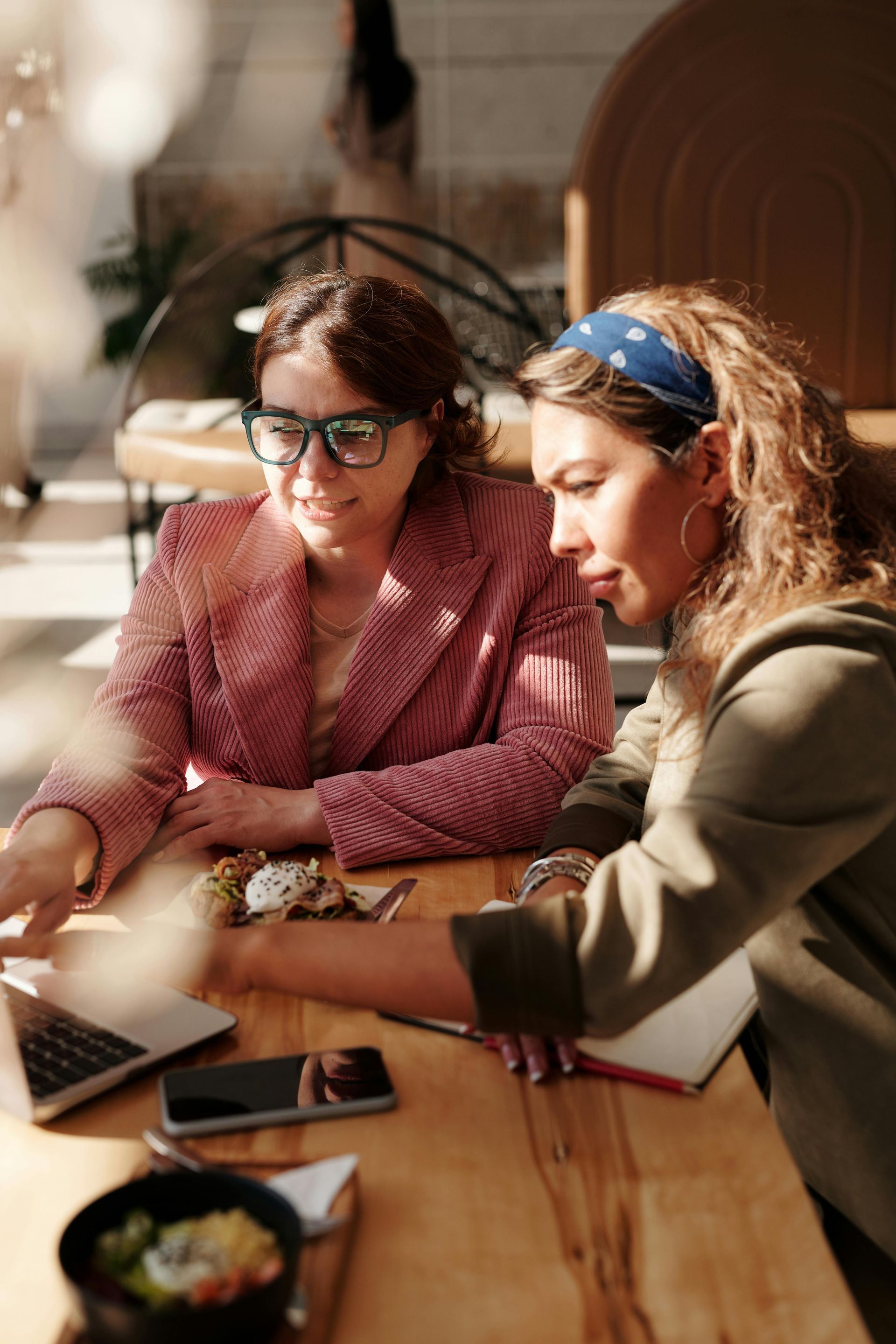 Two women at a table looking at a laptop, one pointing at the screen. Bright, casual setting.