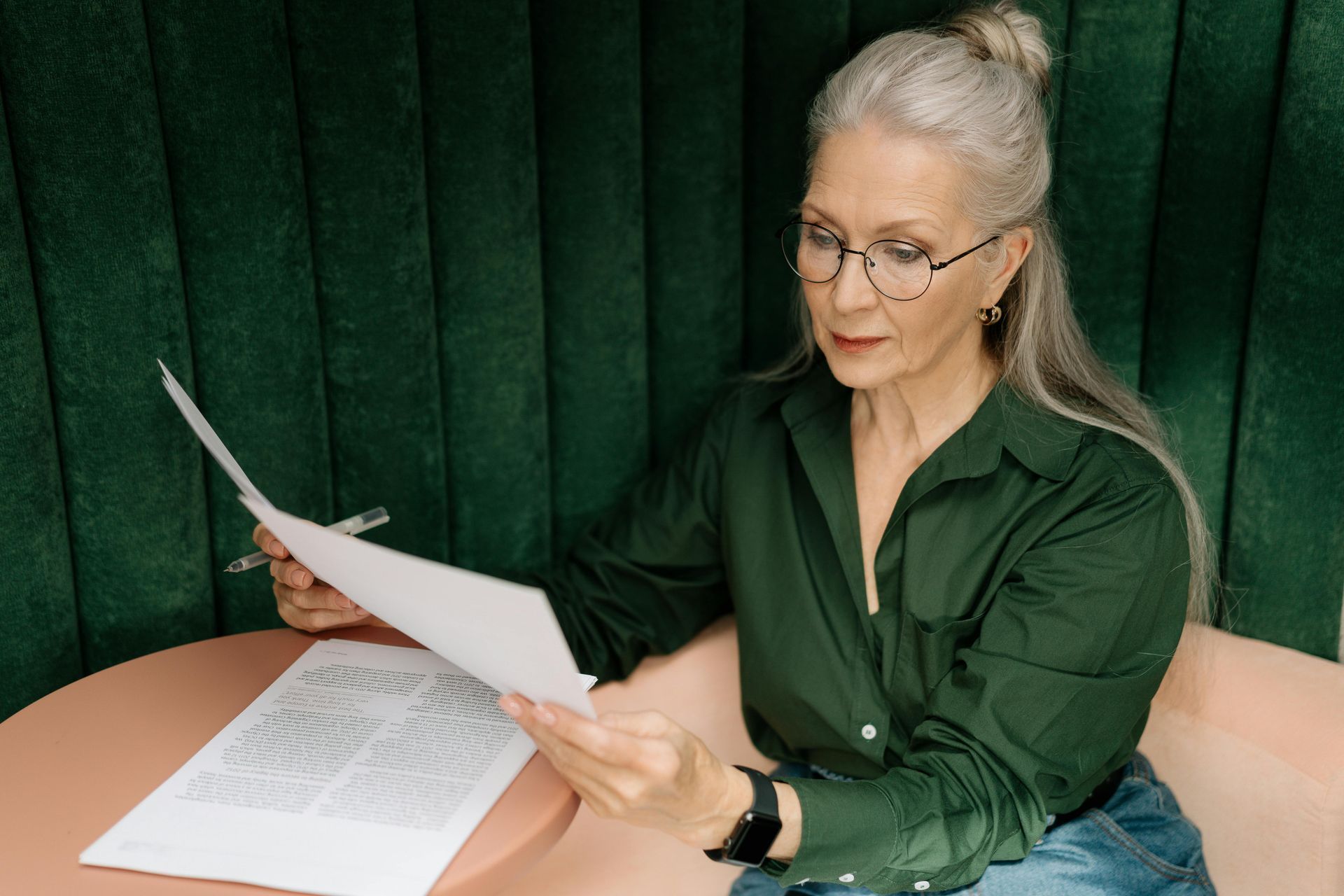 Woman with glasses reviews paperwork at a table; she wears a green shirt and sits in front of a green backdrop.