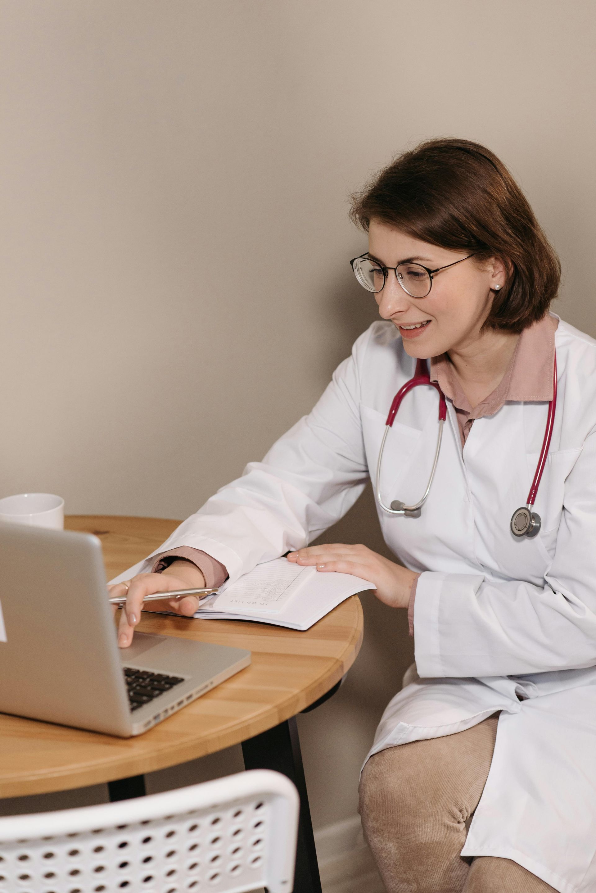 Female doctor smiles while working on a laptop at a wooden table, stethoscope around her neck.