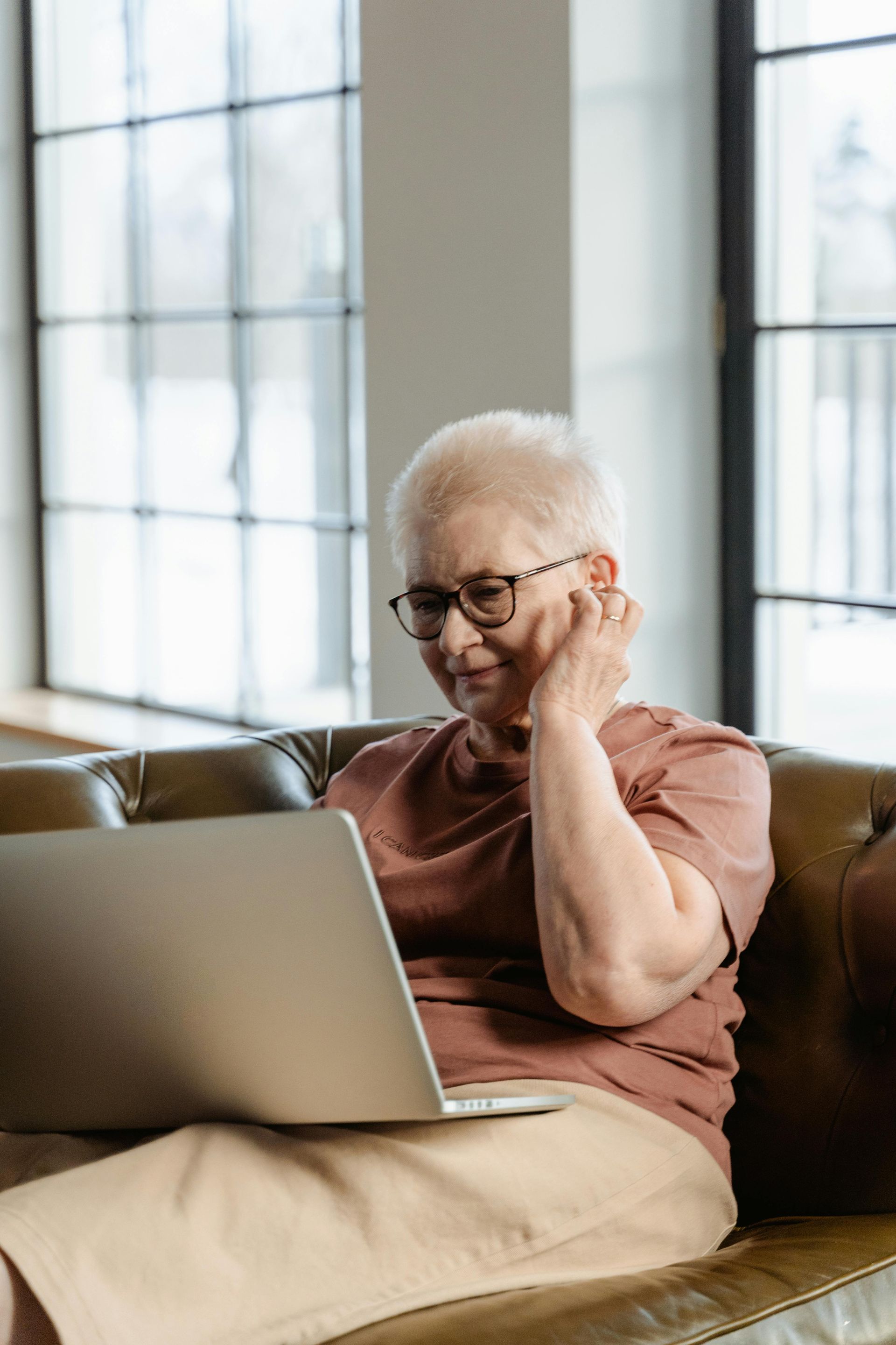 Elderly woman with glasses uses a laptop while sitting on a sofa in a bright room.