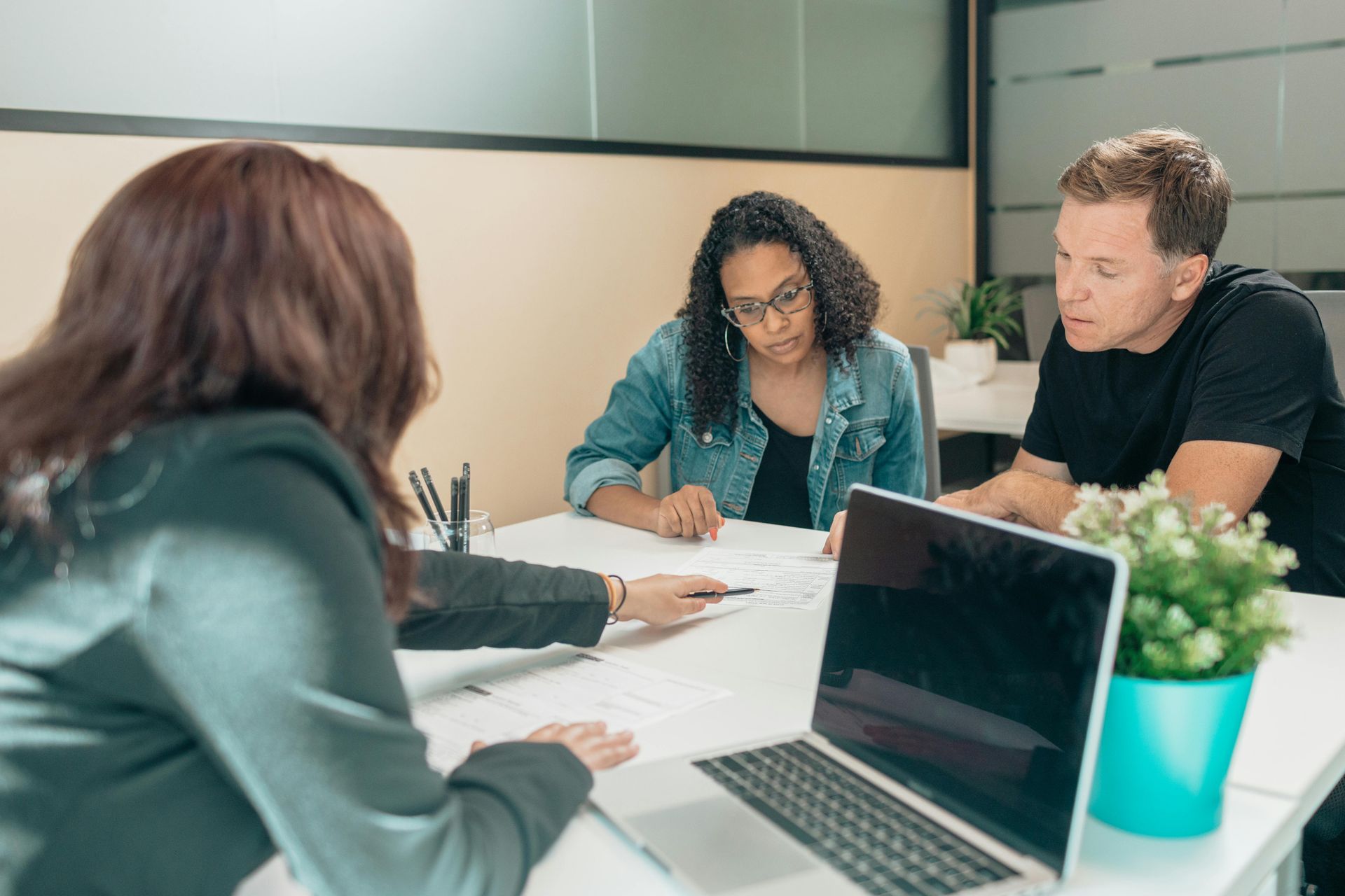 A woman points to documents, assisting a couple at a table with a laptop and small plant.