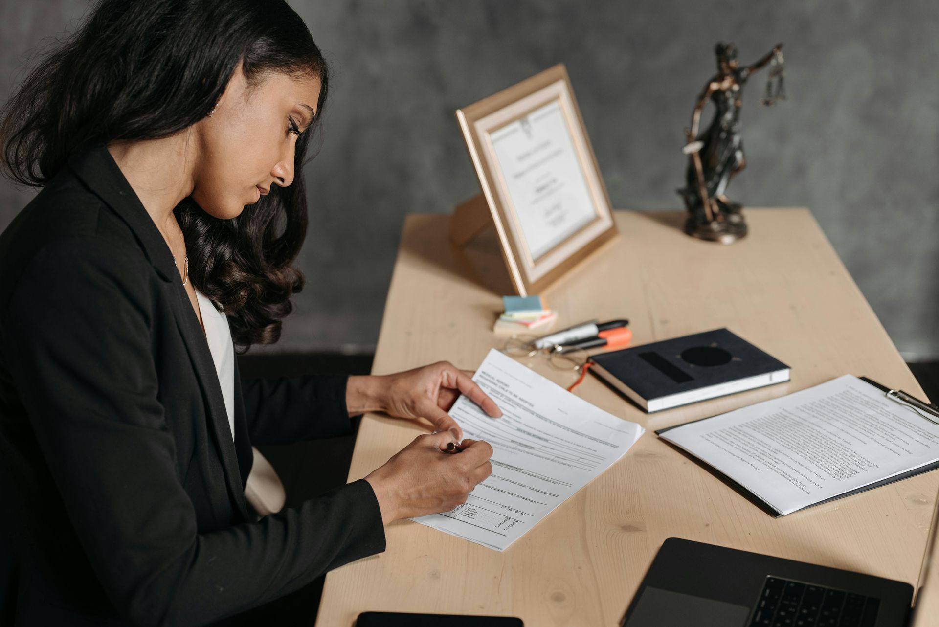 Woman in blazer reviews documents at a desk; statue of justice, diploma, and notebook nearby.