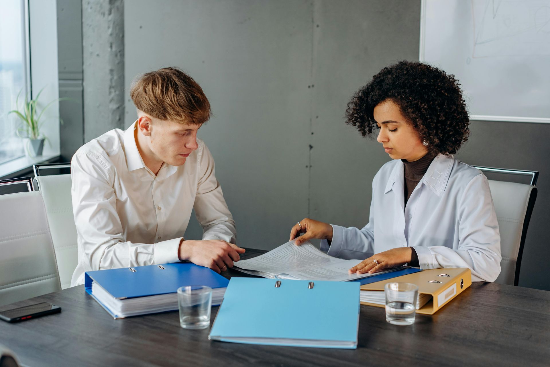 Man and woman reviewing documents at a desk, folders, water glasses, and window in the background.
