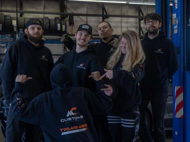 Six people in black hoodies pose at an auto shop. Some point at each other. The shop has tires, tools, and lifts.