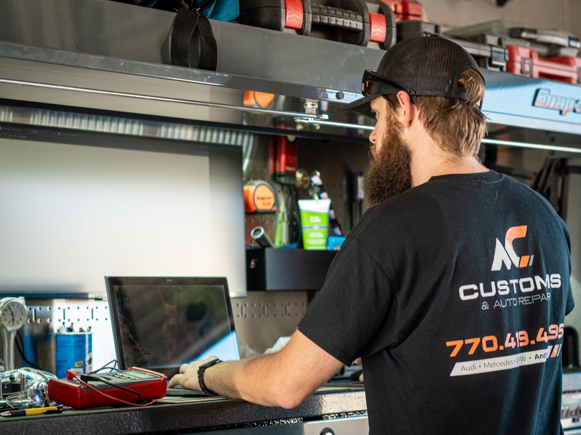 Mechanic working on a laptop in a shop, wearing a black AC Customs shirt.
