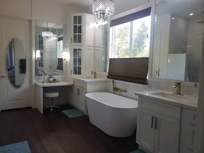 White bathroom with a soaking tub, two sinks, cabinetry, a window, and dark wood floors.