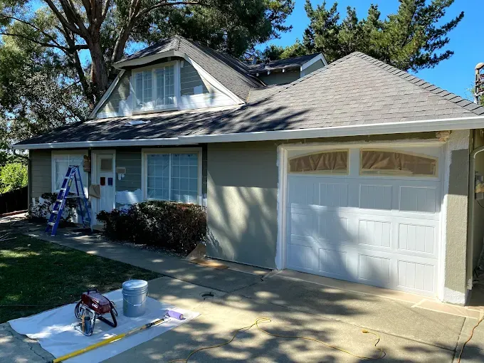 House exterior being painted; white garage door, light green walls, blue ladder, and paint supplies.