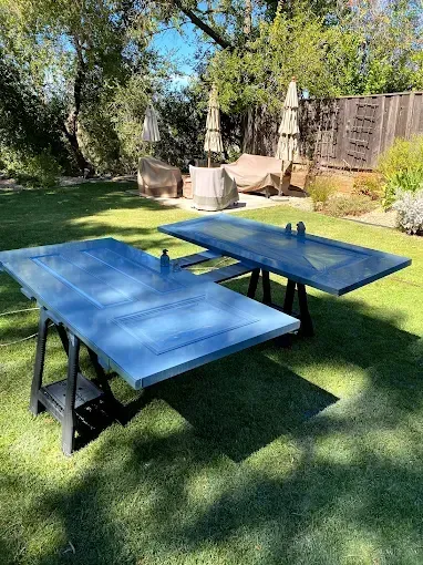 Two blue painted doors on black sawhorses, set on green grass in a backyard.