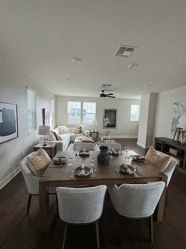 Dining room with table set for dinner; living room visible in background, neutral tones.