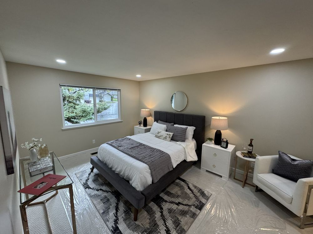 Bedroom with bed, nightstands, rug, and chair. Gray, white, and neutral tones. Window with view of greenery.