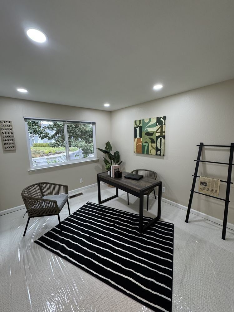 Home office with a desk, two chairs, rug, and artwork. Room has a window and a ladder.