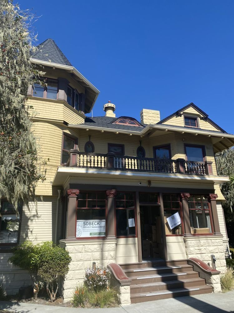 A multi-story Victorian house with a turret, dark roof, light yellow siding, and brown trim against a blue sky.