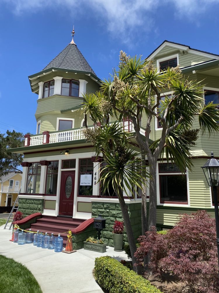 Green Victorian house with turret, red door, and landscaping under a blue sky.