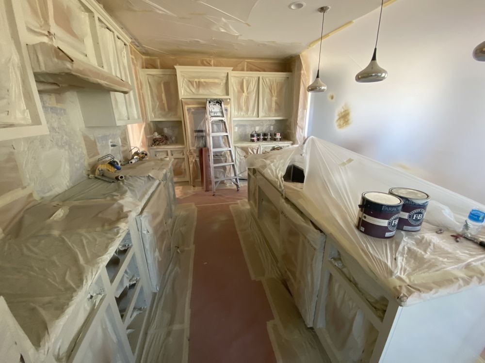 Kitchen being painted, surfaces covered in plastic sheeting. Paint cans and a ladder are visible.