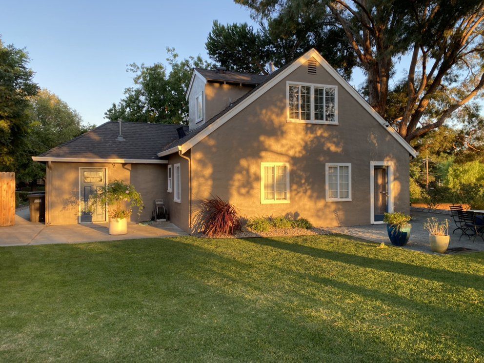 Tan house with gable roof, windows, and door, surrounded by green lawn and trees, lit by golden sunlight.