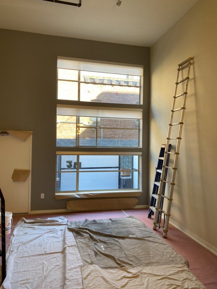 Room with tall windows, ladder, and drop cloth. Walls are gray and beige.