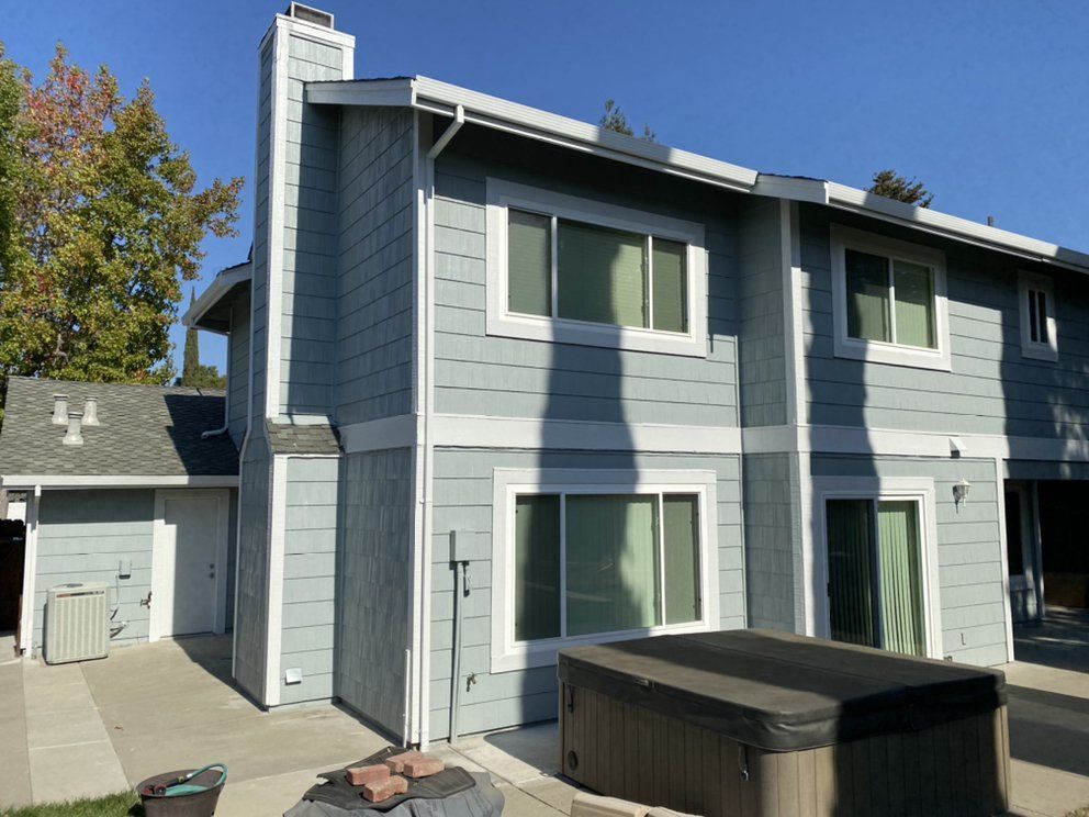 Two-story light blue house with white trim, a chimney, and a hot tub.