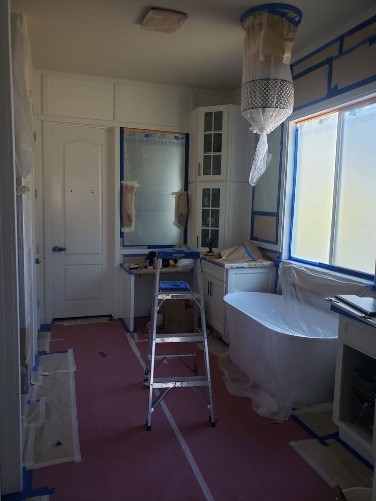 Bathroom interior being renovated; masking tape, drop cloths, ladder present; white cabinets, tub, window.