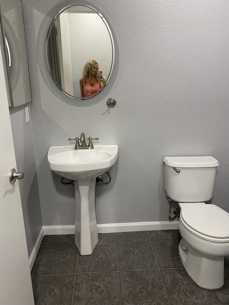 Bathroom with white pedestal sink, toilet, oval mirror reflecting a person, gray walls, and dark flooring.