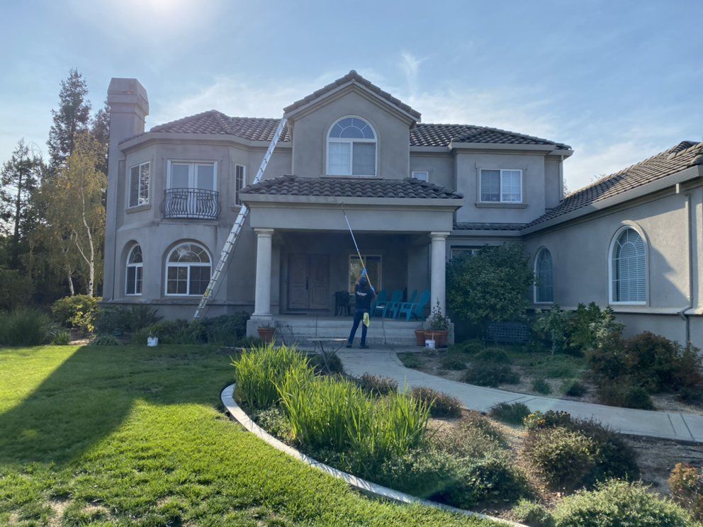 Person washing windows on a large, two-story house with a long pole and ladder. Sunny day.