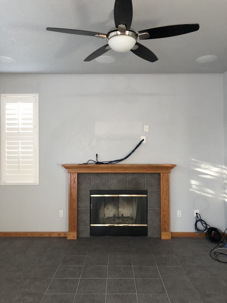 Living room with fireplace, ceiling fan, and window with shutters. Gray walls and dark tiled floor.
