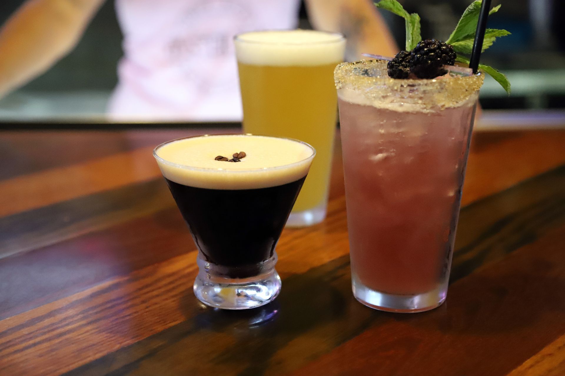 Three colorful cocktails on a wooden bar; a dark, foamy drink, a yellow beer, and a purple drink with blackberries.