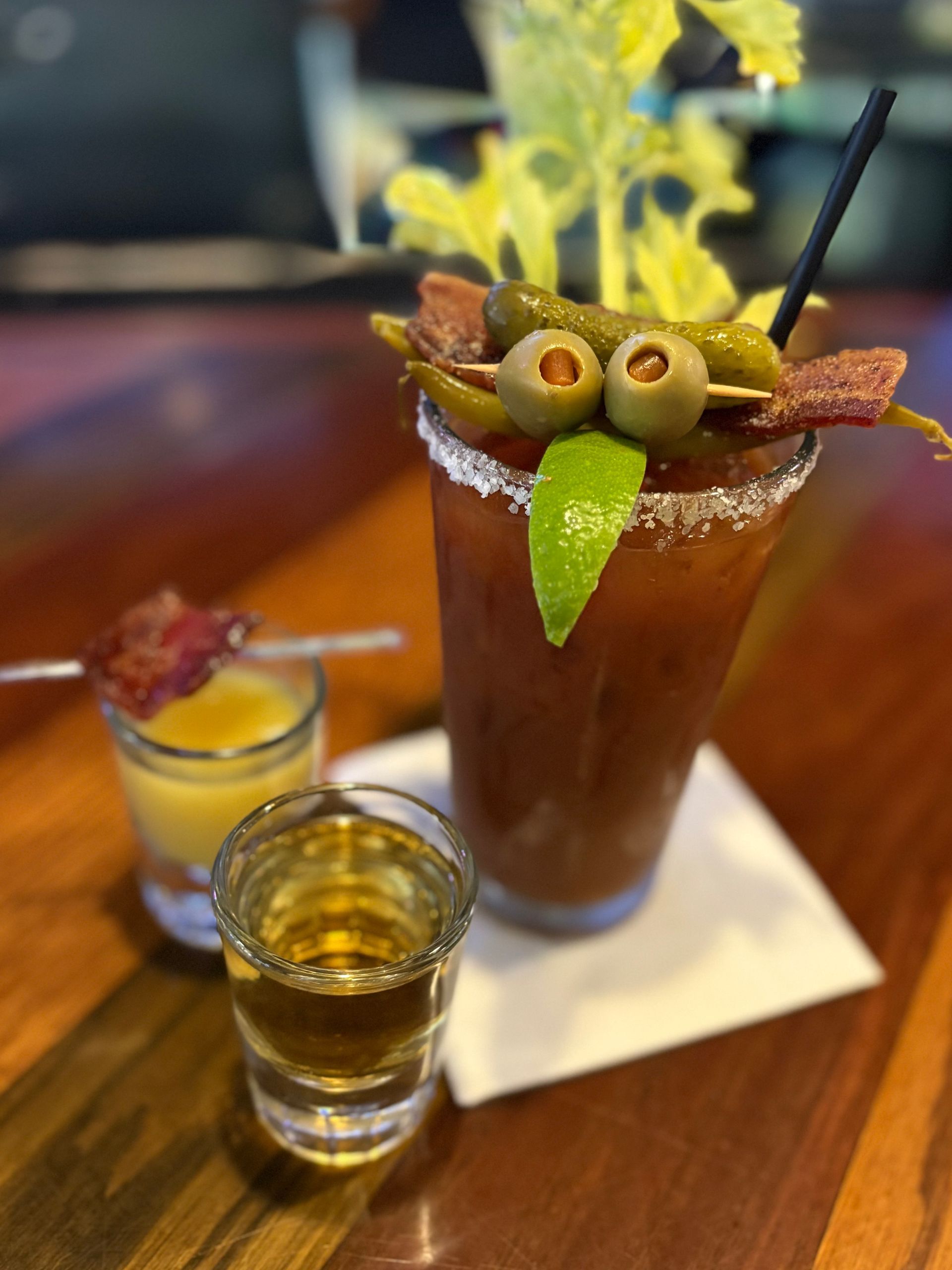Bloody Mary cocktail with garnish, lime, celery, olives, and shots of liquor, on a bar.