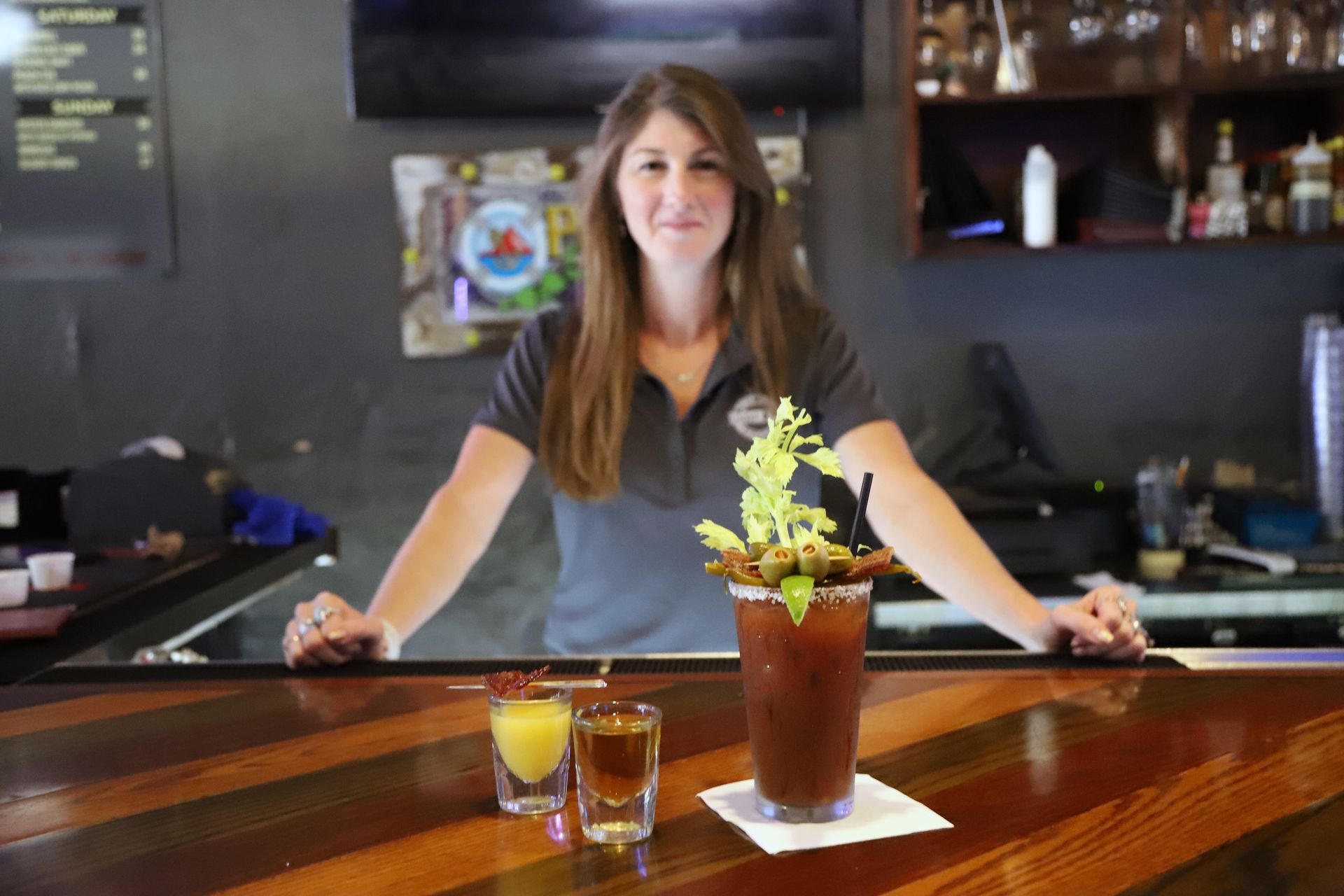 Bartender behind bar with Bloody Mary and shot, arms resting on the counter.