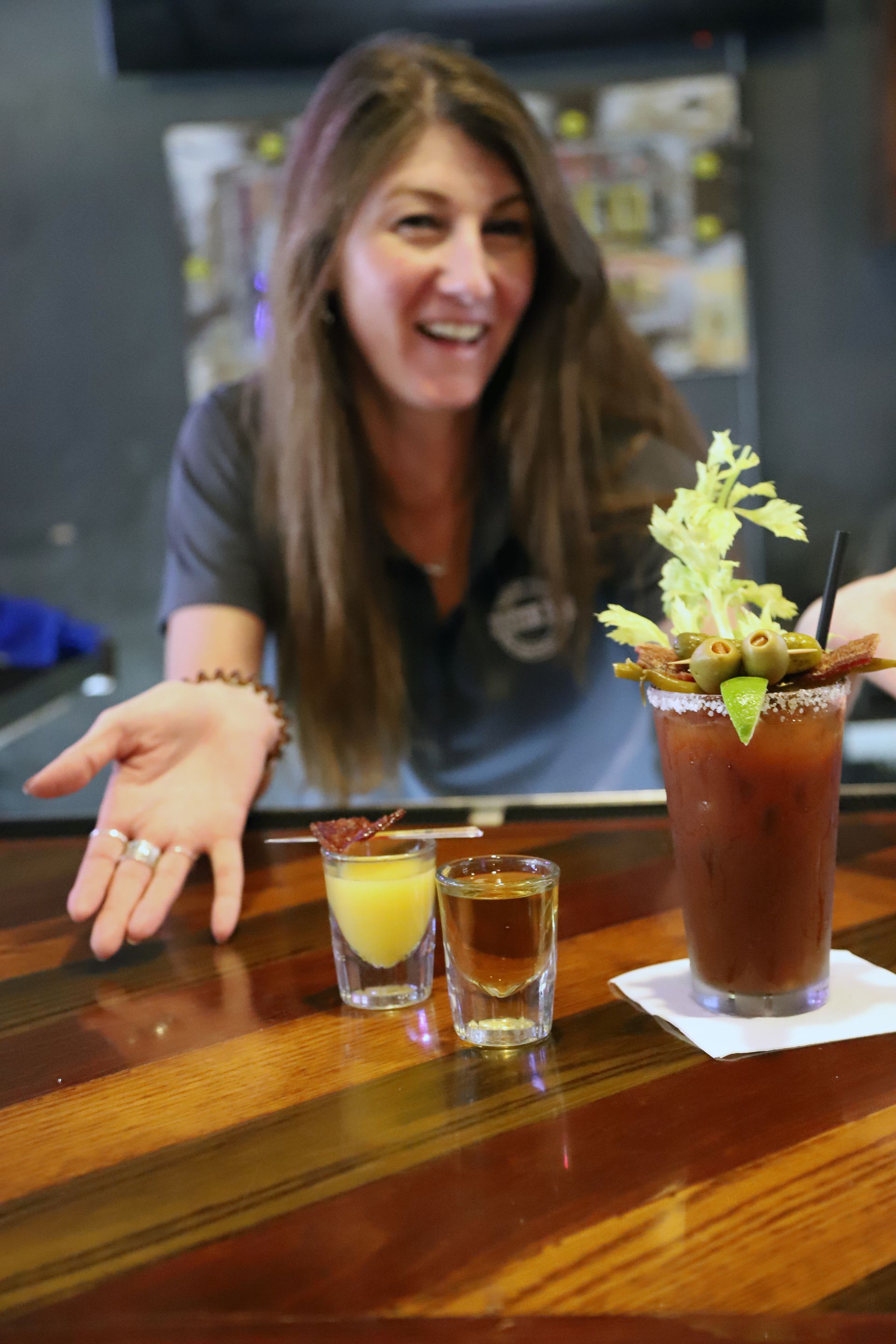 Woman behind bar with Bloody Mary, shots, gesturing to drinks.