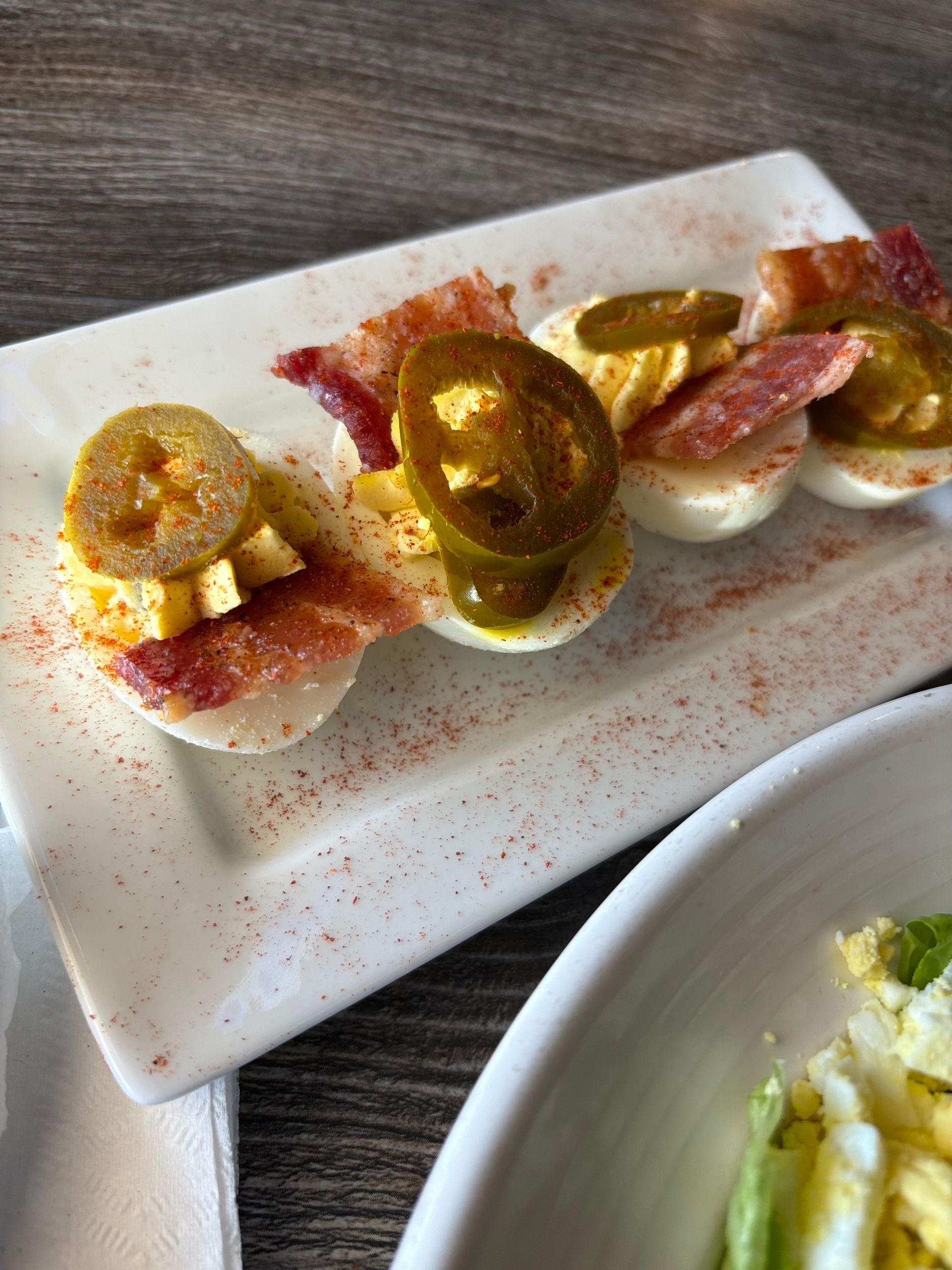 Deviled eggs topped with bacon, pickled jalapeños, and paprika, on a white plate.