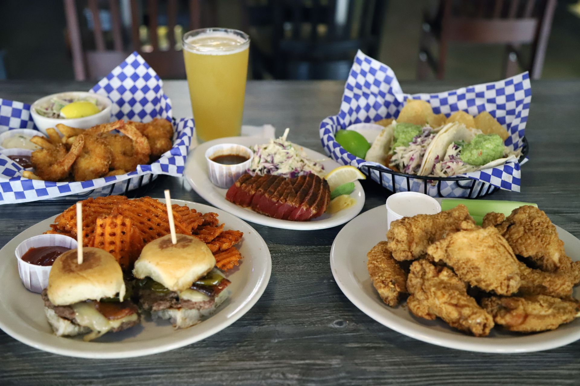 Assortment of pub food: fish and chips, tacos, sliders, wings, steak, beer and sides on a wooden table.