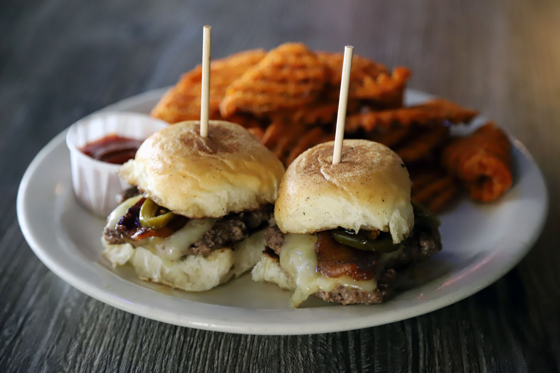 Two sliders with toppings, toothpicks, waffle fries, and ketchup on a white plate.