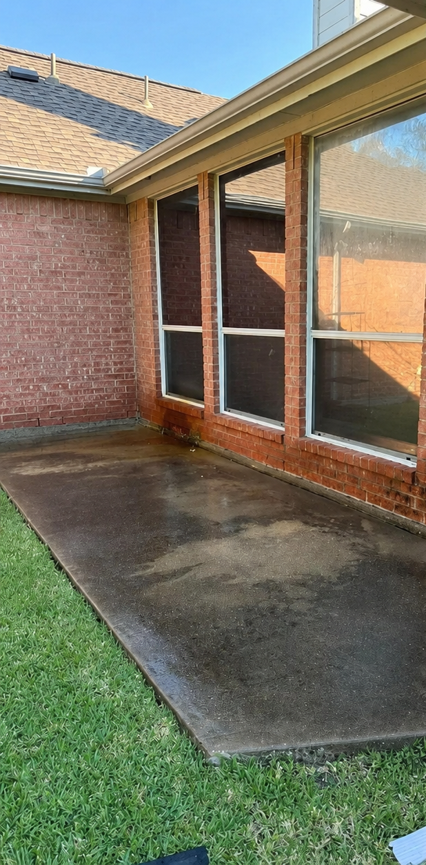 Covered brick patio beside a house, with glass windows and a patch of grass nearby.