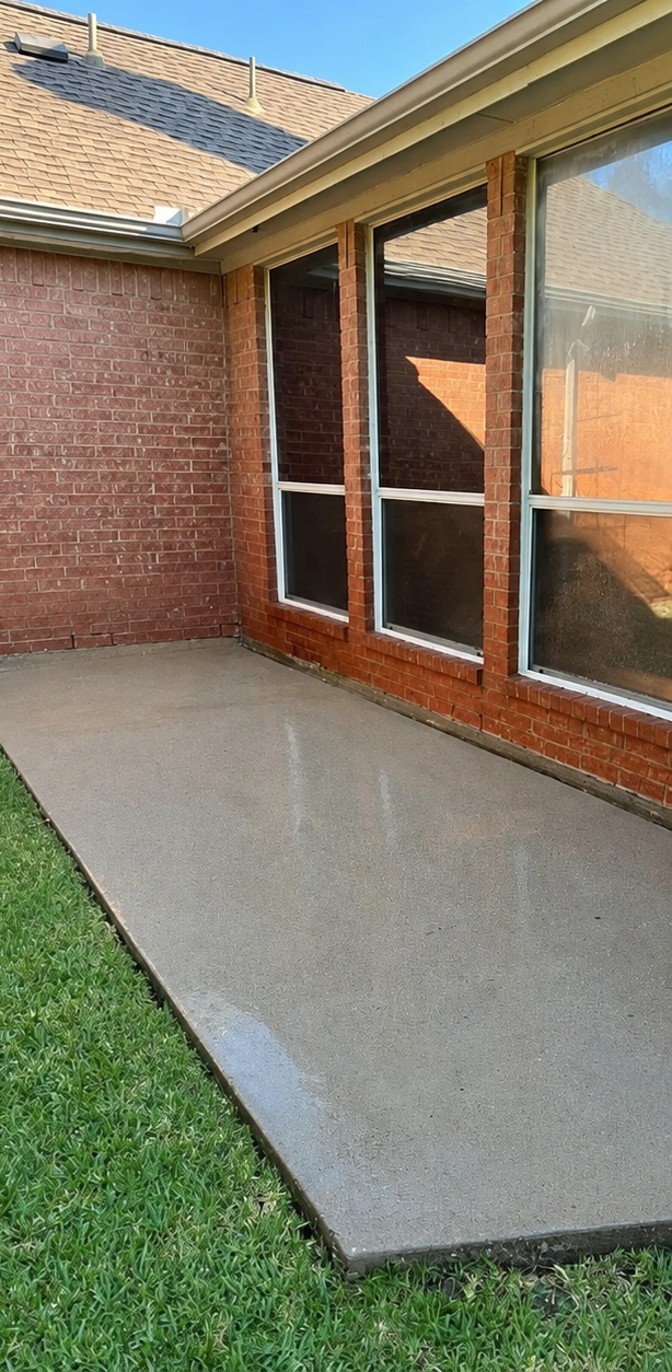 Back patio with concrete slab beside brick house and large windows, bordered by grass