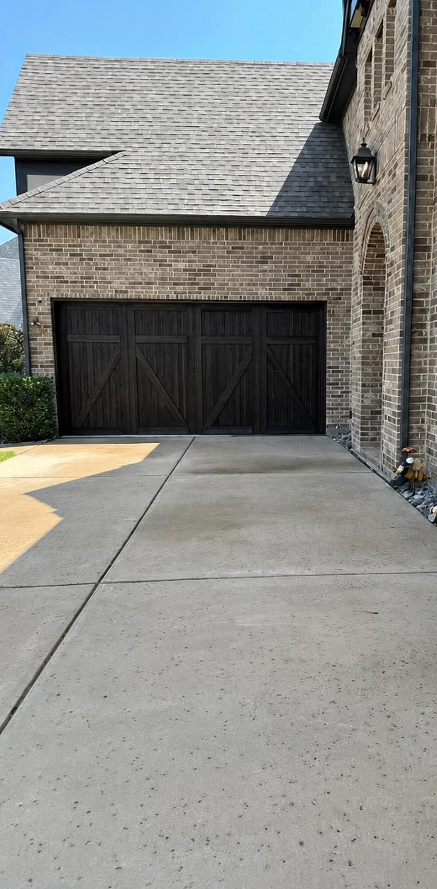 Brick house driveway leading to a dark wooden garage door under a gray shingled roof.