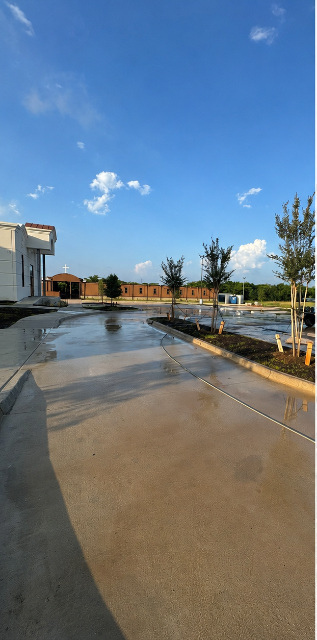 Wet road beside a building under a blue sky, with trees, fencing, and puddles reflecting sunlight
