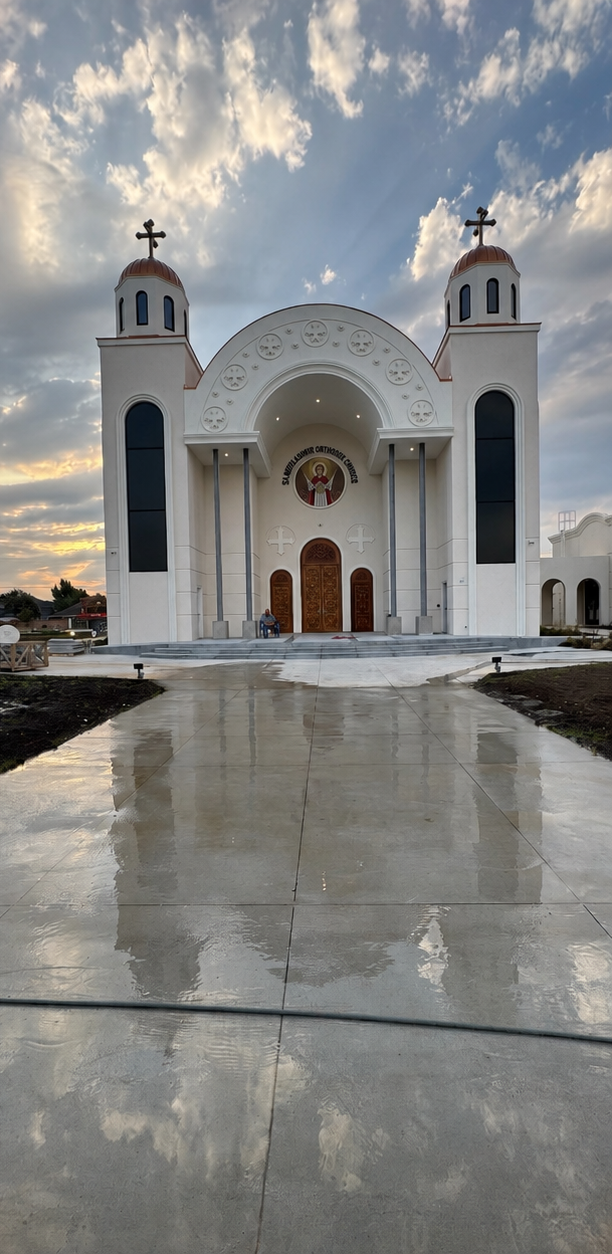 White church with twin bell towers and crosses, viewed from a wet concrete path at sunset.
