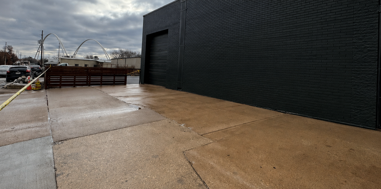 Wet concrete walkway beside a large black building on a rainy day, with a fenced area in the distance