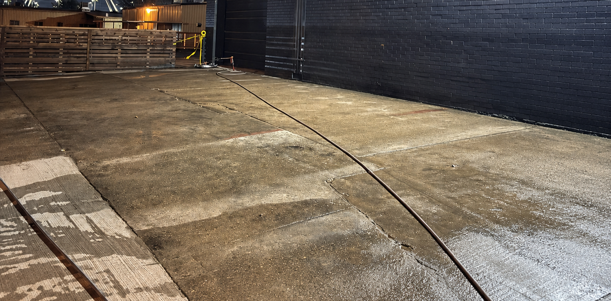 Empty wet concrete courtyard beside a dark wall at night, with lights and stacked pallets in the distance