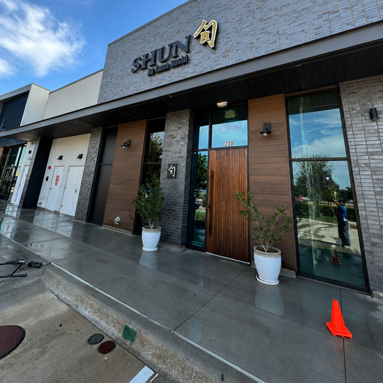 Modern storefront with glass doors, wood accents, potted plants, and wet sidewalk under a blue sky