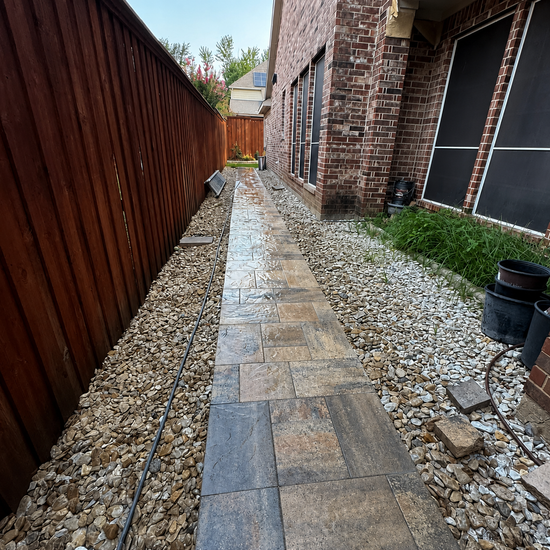Narrow stone walkway beside a wooden fence and brick house, with gravel edging and plants.