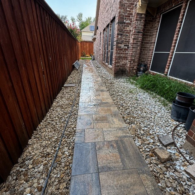 Narrow stone walkway beside a wooden fence and brick house, with gravel edging and plants.