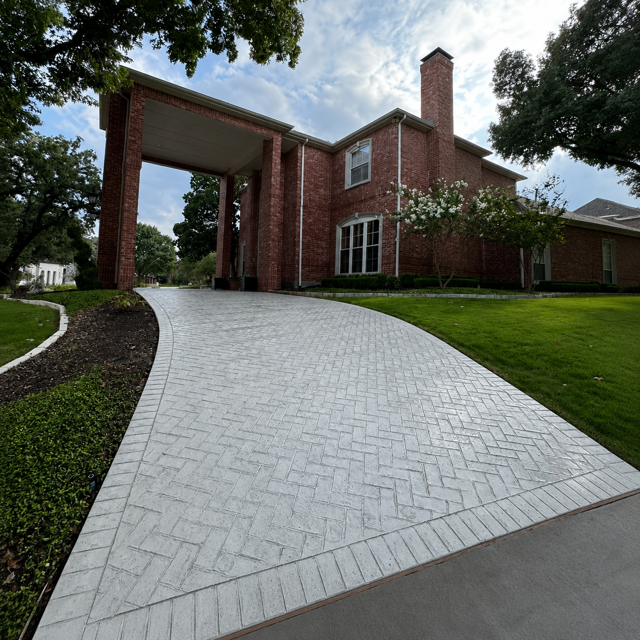 Brick house with covered entry and curved white driveway bordered by green lawn and trees