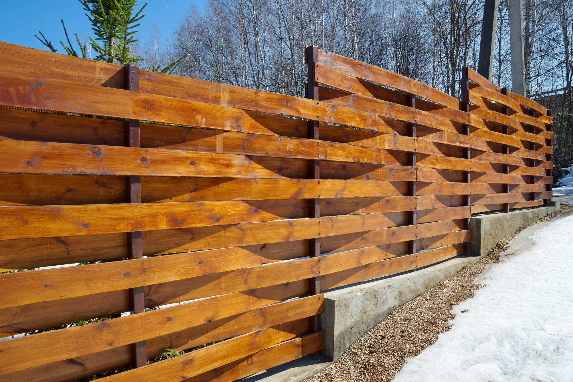 Wooden slat fence with orange stain beside a snowy path and bare trees