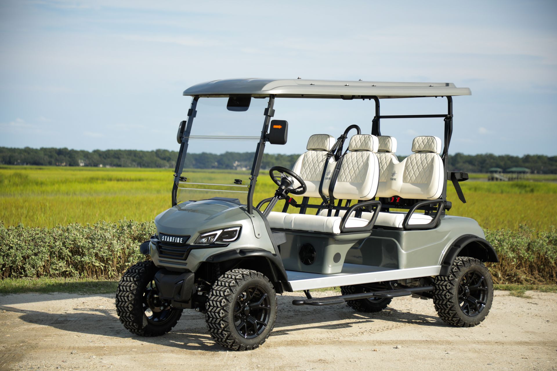 Gray golf cart with lifted suspension and black wheels, parked on gravel beside a field.