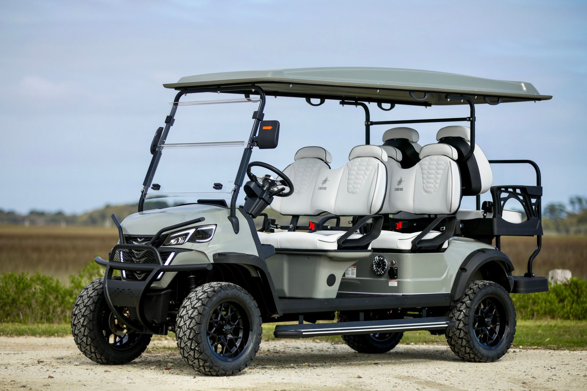 Gray lifted golf cart with white seats, black wheels, and a metal front bumper, parked on a dirt road near water.