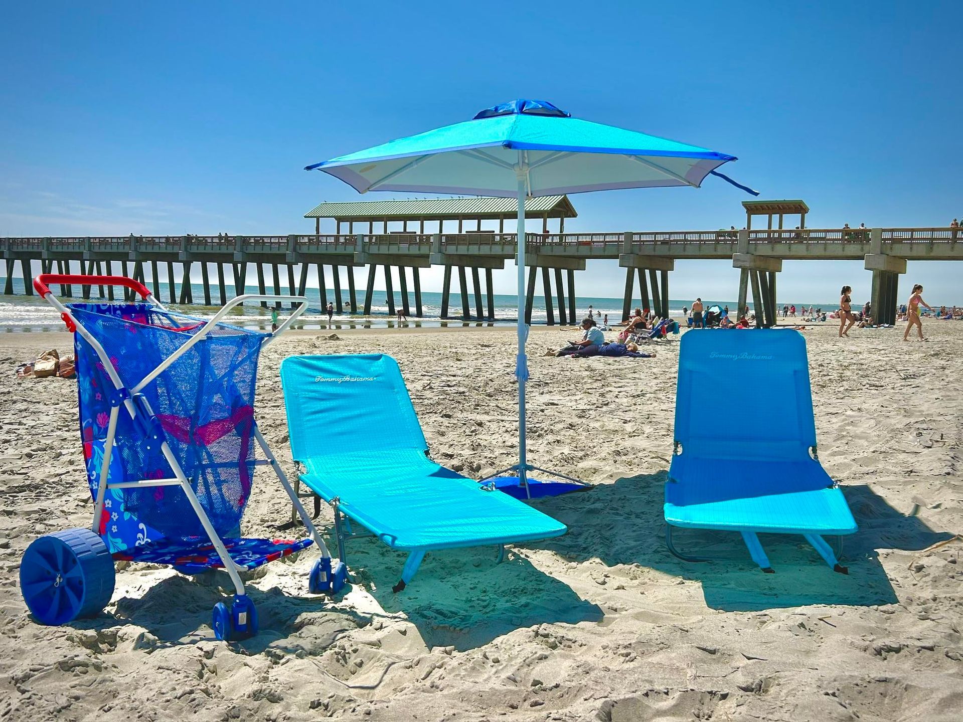 Beach scene: Blue beach chairs, umbrella, and cart on sandy beach, with pier and people in background.