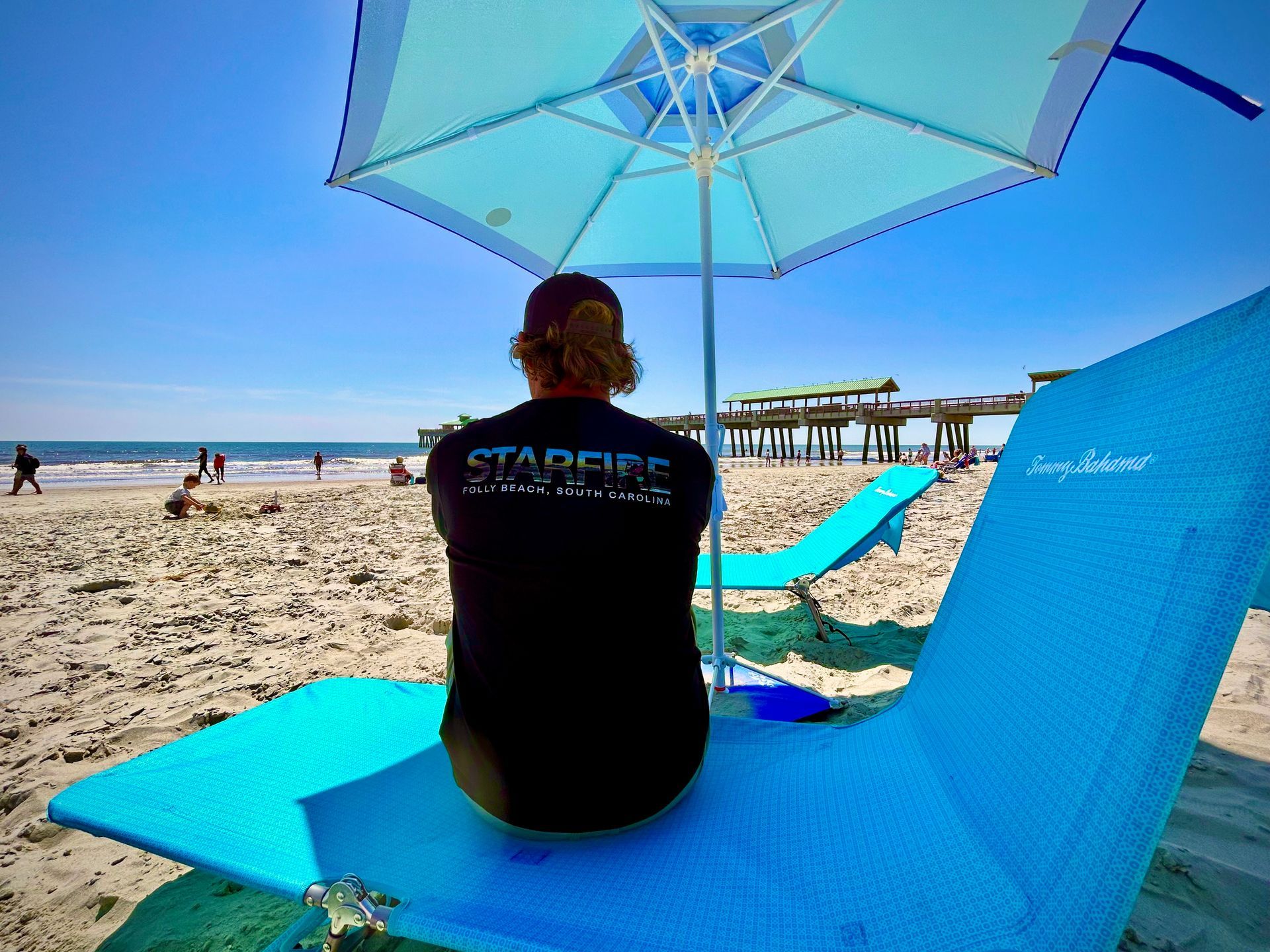 Person in a black t-shirt sitting in a blue beach chair under a blue umbrella, looking out at the ocean on a sunny day.