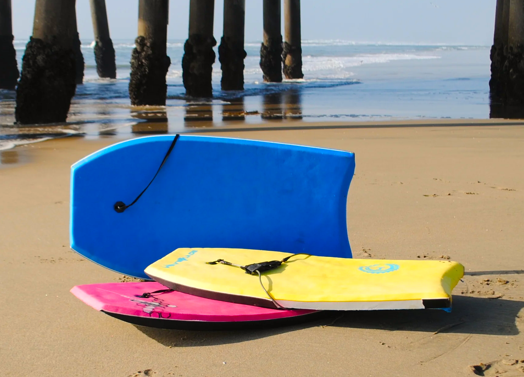 Three colorful bodyboards on the beach; blue, yellow, and pink, with the ocean and pier in the background.