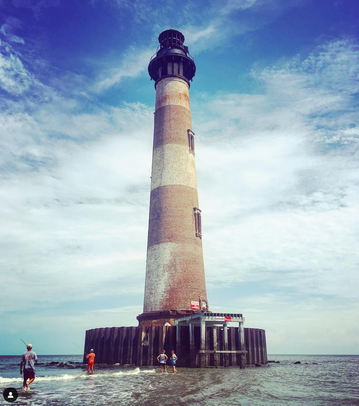 Tall brick lighthouse with brown and white stripes, standing in shallow water under a cloudy blue sky.